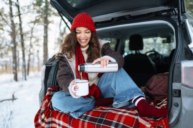 Genç kadın bir termos tutuyor ve güneşli kış ormanlarında arabanın bagajında çay içiyor. Dinlenme, rahatlama, seyahat, yaşam tarzı konsepti.