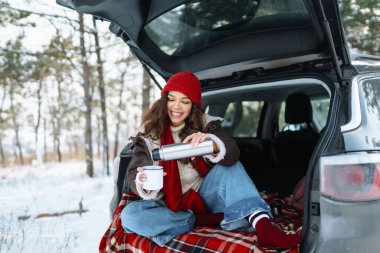 Genç kadın bir termos tutuyor ve güneşli kış ormanlarında arabanın bagajında çay içiyor. Dinlenme, rahatlama, seyahat, yaşam tarzı konsepti.