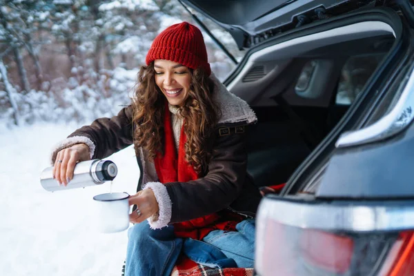 在阳光充足的冬季森林里 年轻的女人拿着一个热水瓶 坐在汽车行李箱里喝茶 生活方式概念 — 图库照片