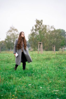 Paltolu genç esmer kadın gündüz vakti yeşil parkta yürüyor.