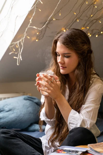 Jovem mulher sentada na cama sonhando e bebendo água quente ou chá em casa — Fotografia de Stock