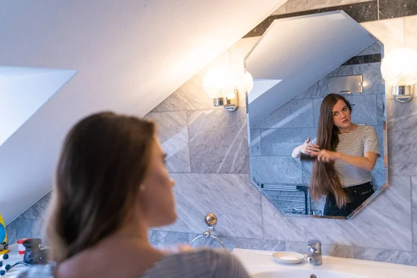 Giovane donna bruna raddrizza i capelli con una spazzola in bagno vicino allo specchio — Foto Stock