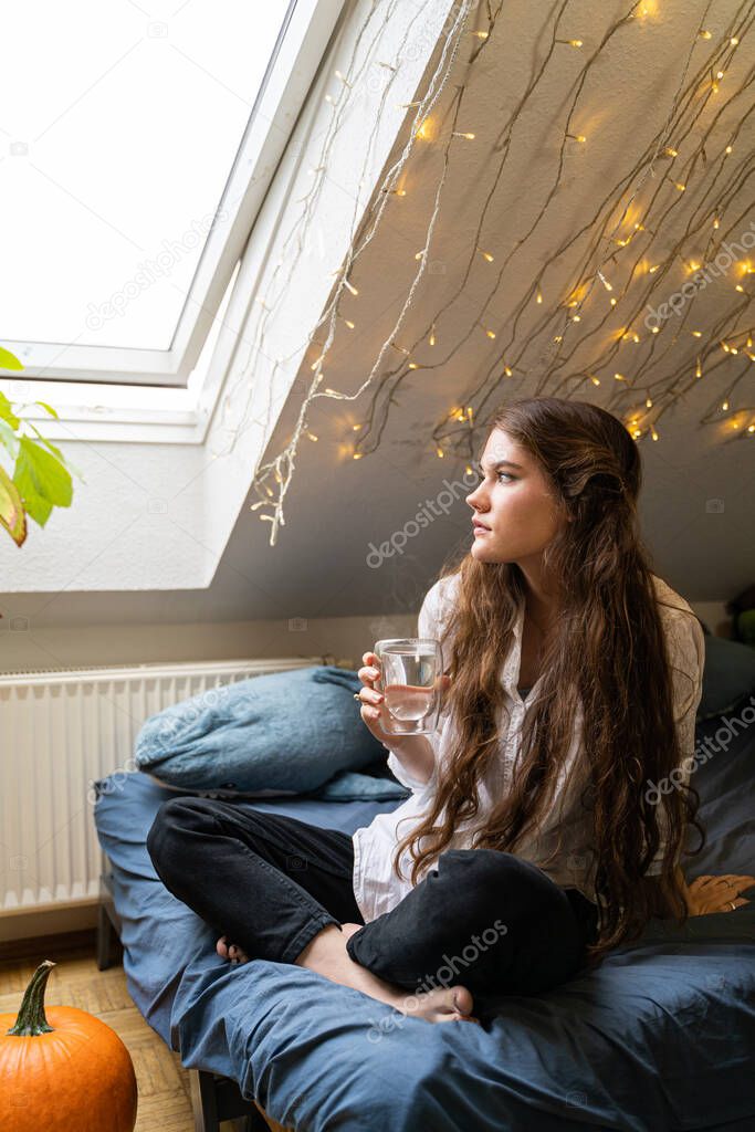 Young woman sitting on bed dreaming and drinking hot water or tea at home