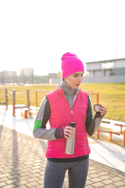 Mujer joven en forma en ropa de abrigo sosteniendo termos metálicos bebiendo té caliente o café — Foto de Stock