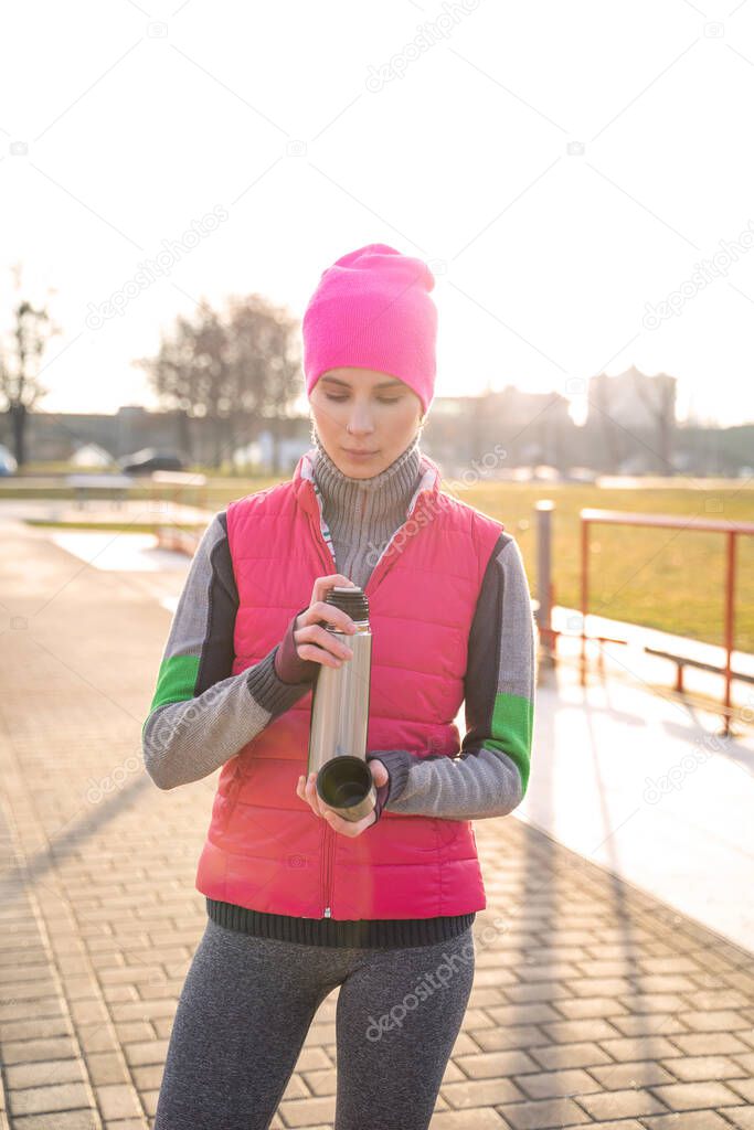 Young fit woman in warm clothes pours hot tea or coffee into mug from thermos
