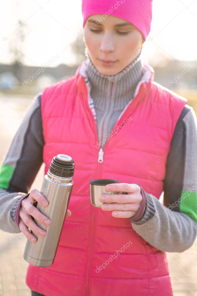 Young fit woman in warm clothes pours hot tea or coffee into mug from thermos