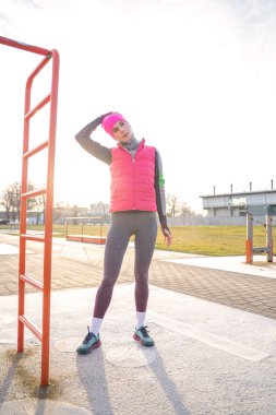 Sıcak giyimli genç bir kadın spor sahasında boynunu uzatıyor.