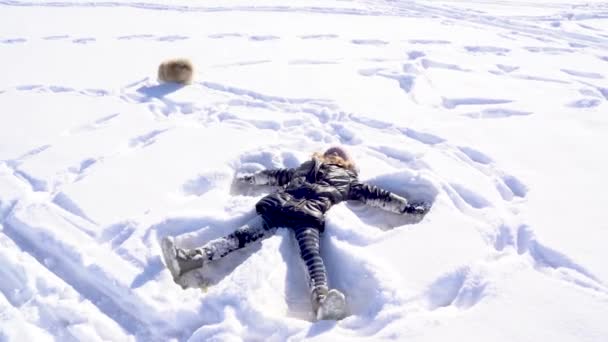 Klein meisje in warme kleren liggend op sneeuw doen sneeuw engel met hond huisdier Spitz — Stockvideo