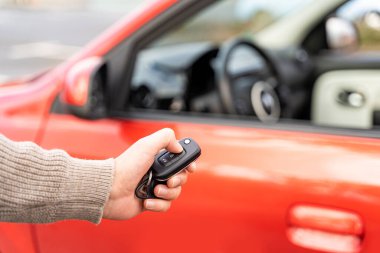 Male hand holding electronic remote key pushing button near red rental car