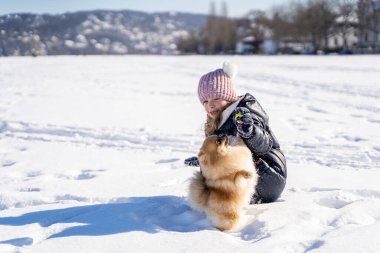 Kalın elbiseli küçük bir kız kar üstünde köpek Spitz 'e yemek veriyor.