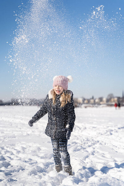 Молодая улыбающаяся дошкольница играет со снегом на улице в солнечную зимнюю погоду