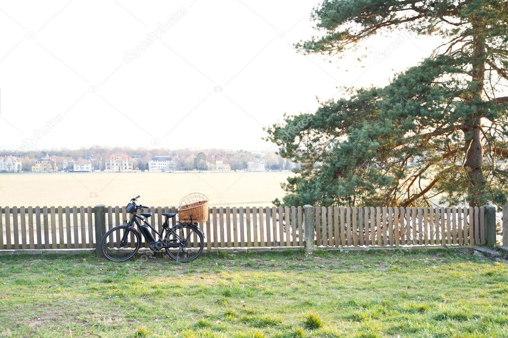 Modern electric bike with wicker basket parked near wooden fence on meadow
