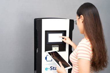 Genç Asyalı Taylandlı, Vietnamlı ya da tişörtlü Çinli kadın, beyaz tablet tutan kot pantolon ve gözlüklü, dışarıda ya da içeride dokunmatik ekran elektrikli araba şarj istasyonu kuruyor. Araba paylaşımı, modern teknoloji