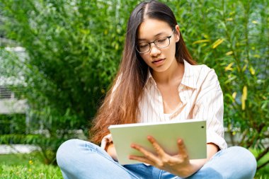 Genç Asyalı Taylandlı, Vietnamlı ya da Çinli sıradan giyinmiş ve gözlüklü bir kadın çalılığın yanında yeşil çimlerin üzerinde oturmuş web kameralı tablet kullanıyor ya da yaz günü film seyrediyor.. 