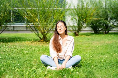 Genç Asyalı Taylandlı, Vietnamlı ya da Çinli rahat rahat oturmuş ve güneşli yaz günlerinde çalıların ve ofis binalarının yakınındaki yeşil çimlerin üzerinde hayal kuruyor.. 