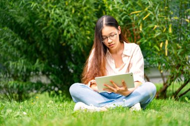 Genç Asyalı Taylandlı, Vietnamlı ya da Çinli sıradan giyinen ve gözlüklü yeşil çimenlerin üzerinde çalıların yakınında oturan güneşli yaz havasında dışarıda modern tablet kullanan, okuyan ya da ödev yapan genç bir kadın.