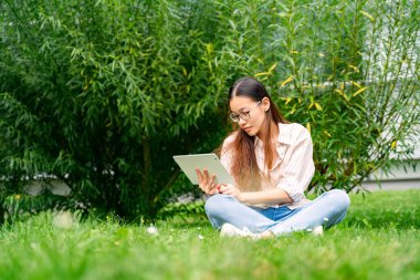 Genç Asyalı Taylandlı, Vietnamlı ya da Çinli, günlük giysiler ve gözlüklerle yeşil çimlerin üzerinde, çalılıkların yakınında oturuyor güneşli yaz gününde parkta beyaz tablet kullanarak çalışıyor, çalışıyor ya da okuyor.