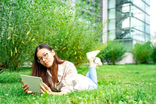 Portrait of young cheerful Asian Thai, Vietnamese or Chinese woman in casual clothes and glasses lying on green grass loan outside near univercity or school working reading or learning using tablet