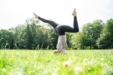 Salamba Sirsasana yoga pozu veren genç beyaz ya da Asyalı zayıf kadın yaz güneşli parkında yeşil çimen kredisinin dengesini sağlamak için geyik bacaklarıyla başucunda duruyor. Etkin yaşam biçimi