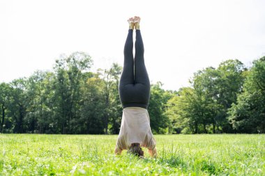 Genç Kafkas ya da Asyalı zayıf kadın yaz güneşli parkında meditasyondan ya da yeşil çimen kredisiyle ilgili pilates yaptıktan sonra Salamba Sirsasana yoga pozu veriyor. Arka plan, yüz yok.