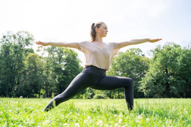 Spor kıyafetleri içindeki beyaz tenli genç kadın yoga pozu veriyor ya da öğleden sonra yeşil ağaçlarla güneşli bir parkta günlük vücut ve sağlık hizmetleri rutini olarak pilates yapıyor. Yan görünüm