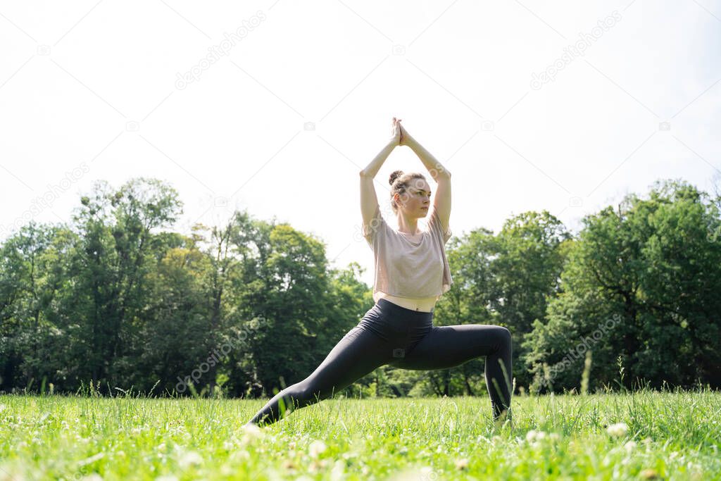 Young caucasian slim fit woman in sportswear doing yoga practice pose or pilates with hands up after wake up, meditation or workout on grass meadow in summer sunny park with green trees on afternoon.