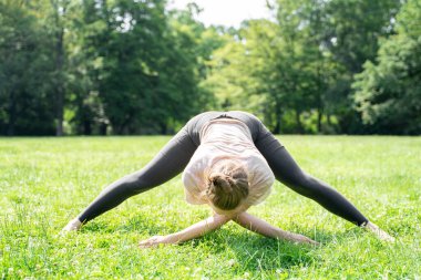 Spor kıyafetleri giyen, aerobik yapan, spor yaptıktan sonra ellerini yerde çapraz yapan ya da öğleden sonra güneşli bir parkta yeşil çimlerde pilates yapan beyaz tenli genç bir kadın. Arka plan