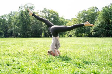 Salamba Sirsasana amuda çalışan beyaz tenli ya da Asyalı sıska kadın başındaki yeşil çimen kredisiyle yaz güneşli parkında yoga pozu veriyor. Sağlıklı bir yaşam tarzı, yan görüş, yüzü yok.