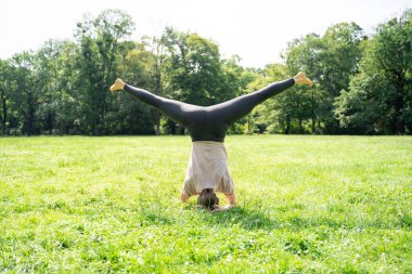 Salamba Sirsasana amuda çalışan beyaz tenli ya da Asyalı sıska kadın başındaki yeşil çimen kredisiyle yaz güneşli parkında yoga pozu veriyor. Sağlıklı bir yaşam tarzı, arka plan, yüzü yok.