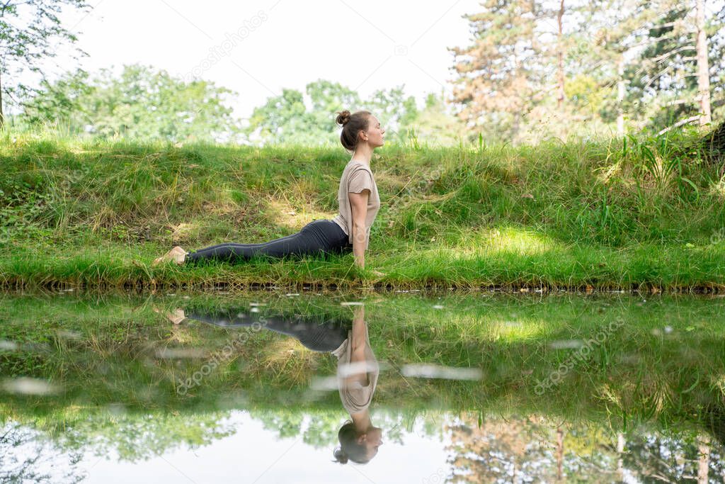 Young caucasian fit woman in sportswear doing sun salutation yoga practice or back stretching after meditation, workout or pilates on green grass near calm river in summer park. Active lifestyle