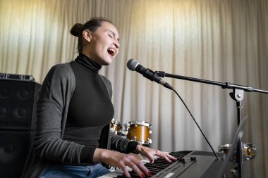 Genç Asyalı ya da Kafkasyalı esmer kadın, müzik stüdyosunda, okulda, etkinlikte ya da konser sahnesinde duygusal olarak yüksek sesle şarkı söylüyor ve dijital klavye elektronik piyanosu çalıyor.