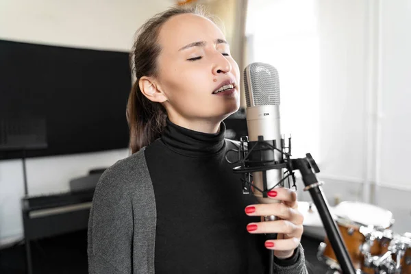 Young Asian or Caucasian brunette woman emotionally singing lyric song to microphone with closed eyes alone in music studio. Practicing and learning, preparing for exam or lesson in conservatory