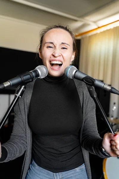 Young Japanese or Caucasian brunette woman having fun fooling around singing rock song on two microphones emotionally loud and with pleasure alone in music studio or school, event or concert stage.