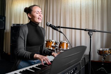 Genç Asyalı ya da Avrupalı esmer kadın mikrofon ve yüksek sesle şarkı söyleyerek ve dijital klavye elektronik piyano çalarak müzik stüdyosunda, etkinlikte ya da konser sahnesinde tek başına eğleniyor.