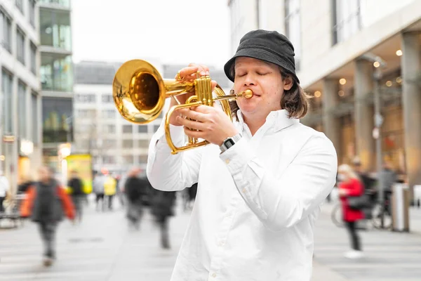 Young caucasian blond man with long hair and closed eyes in white shirt and hat playing jazz on golden trumpet with pleasure, standing in middle of crowded modern downtown street with clothing stores