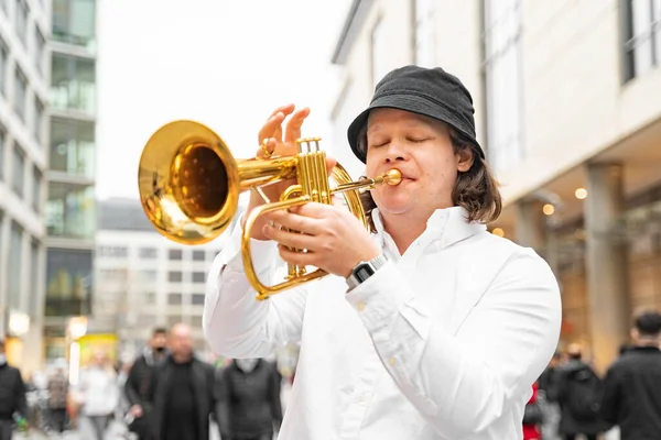 Young caucasian blond man with long hair and closed eyes in white shirt and hat emotionally with feeling playing jazz on golden trumpet with pleasure, in middle of crowded modern street in Dresden