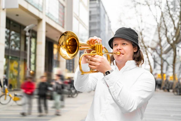 Young caucasian long-haired blond man in white shirt and hat playing funky jazz on golden trumpet with pleasure standing in middle of crowded modern downtown street with department stores, in Germany