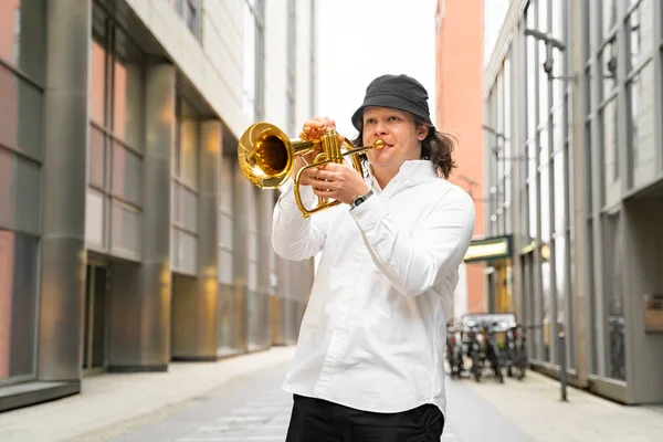 Young caucasian blond cheerful smiling man in white shirt and hat playing funky jazz on golden trumpet with pleasure alone, standing between modern skyscrapers in downtown with clothing stores