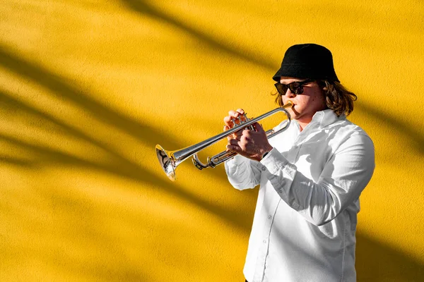 Young caucasian long-haired man in white shirt, hat and sunglasses playing funky jazz on silver trumpet with pleasure standing near yellow wall on city street on concert, event or school exam outside