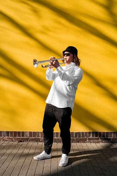 Young caucasian long-haired male musician in white shirt, hat, sunglasses and sneakers playing classic jazz on silver trumpet with pleasure standing near yellow wall on street outside on sunny day