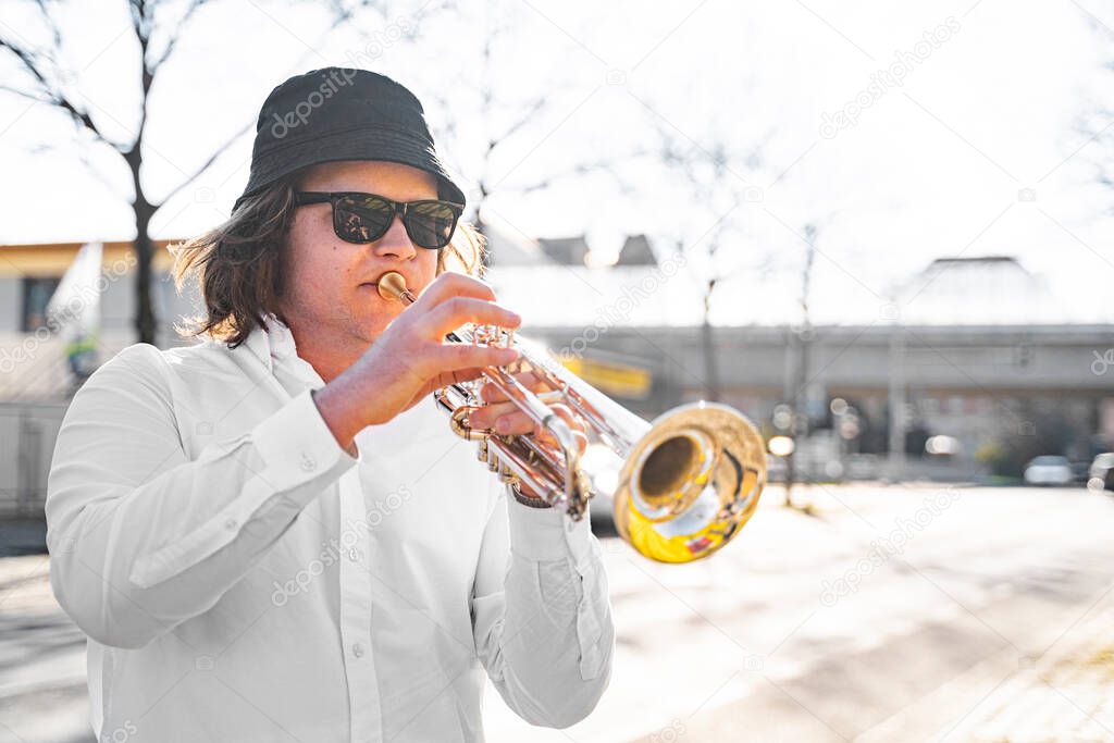 Young European or American male musician jazzman with long hair in white shirt, hat and sunglasses playing on trumpet on sunny town street near road on summer day. Event or concert, masterclass