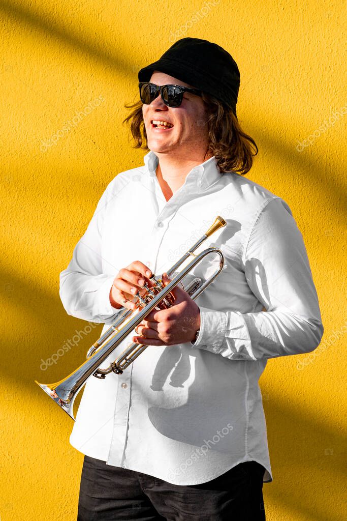 Young caucasian blond cheerful smiling man in white shirt, hat and sunglasses holding silver trumpet standing near yellow wall with tree shadows on sunny street on summer day ready to play on concert