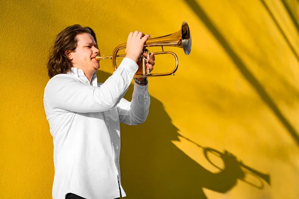 Young caucasian long-haired man in white shirt playing classic or funky jazz on golden trumpet with pleasure standing near yellow wall on city street on concert, event, school exam or festival outside