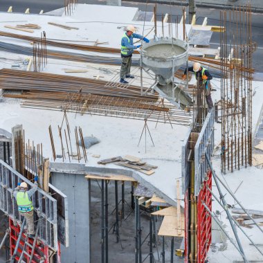 İnşaat alanı mavi gökyüzüne karşı çalışıyor. Hidrolik yelken yelkenli vinçleri ve temele beton dökülen işçileri kandırıyor. İnşaat alanındaki inşaat çalışmalarına çimento dökülüyor. 