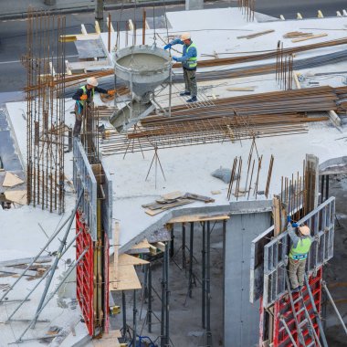 İnşaat alanı mavi gökyüzüne karşı çalışıyor. Hidrolik yelken yelkenli vinçleri ve temele beton dökülen işçileri kandırıyor. İnşaat alanındaki inşaat çalışmalarına çimento dökülüyor. 