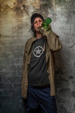 Keş adam yeşil anorak ve mavi eşofmanlı taş duvarın yanında