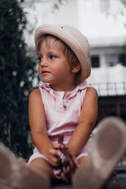 A little girl in a cap with big ears near a tree. A blonde child in a dress looks away. A dreamy mood, raising children, a happy childhood