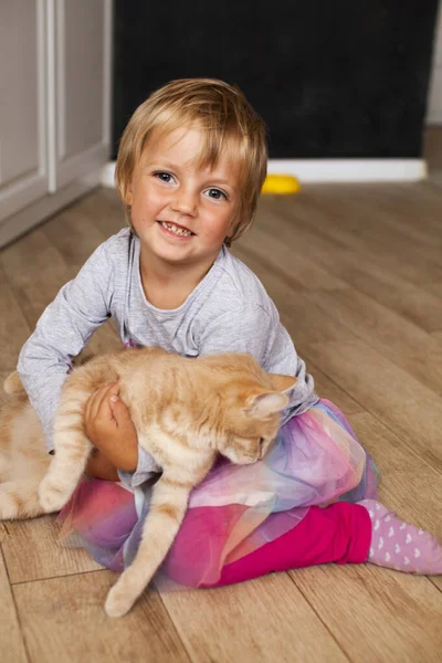 A girl with a red cat in her arms. A smiling blonde girl with short hair plays with a pet at home