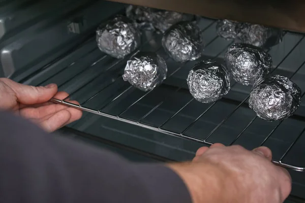 A man puts raw potatoes wrapped in foil on wire rack in the oven. — Fotografia de Stock