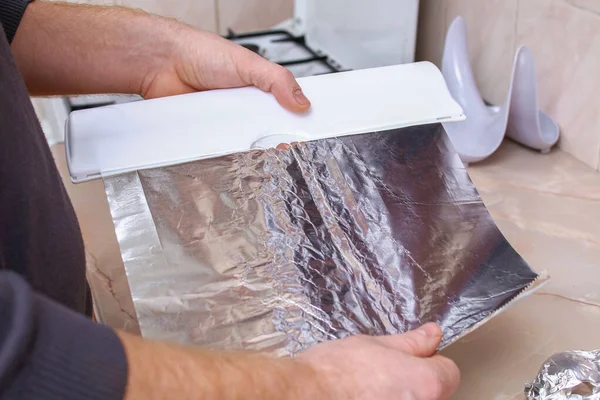 A man tears off the foil from plastic dispenser to wrap potatoes in it and bake them in the oven. — Fotografia de Stock